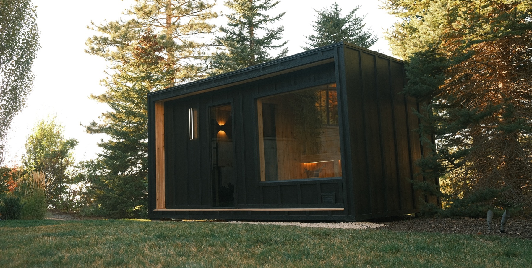 A charred-timber New Primitive sauna set among pines at golden hour
